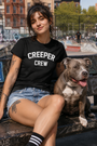 Woman in black "CREEPER CREW" tee, denim shorts, and a gray pit bull on a park bench.
