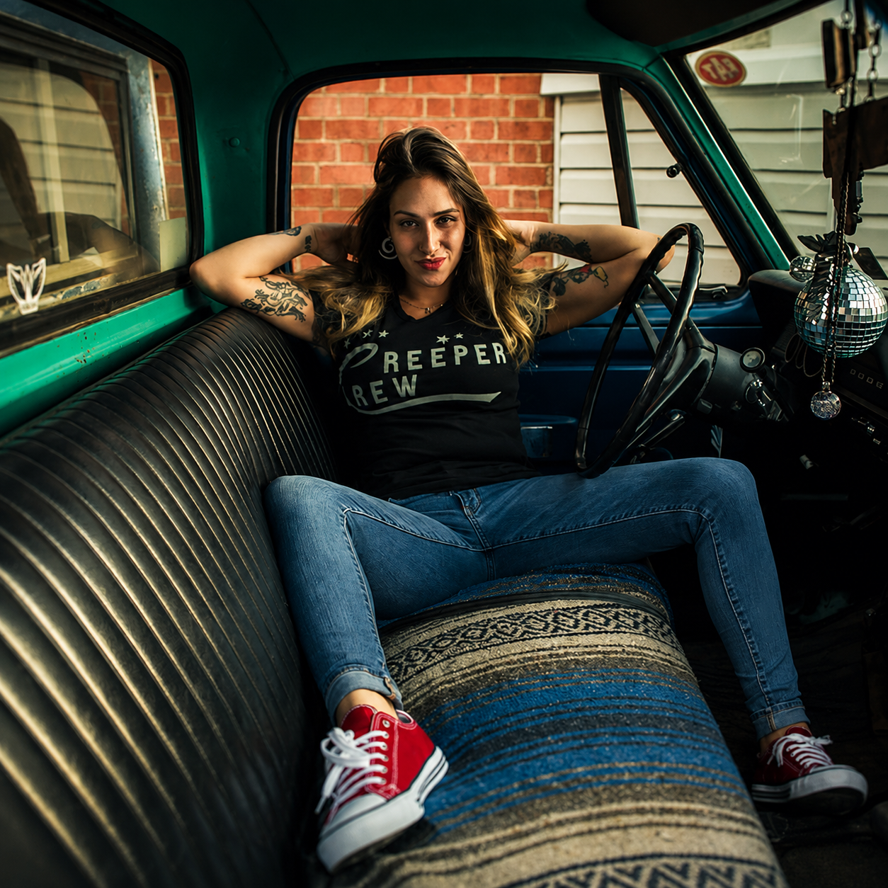 Woman in black SMUTLIFE "CREEPER CREW" tee, blue jeans, and red sneakers sitting in a vintage truck.