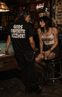 Man in black "GODS FAVORITE AZZHOE" t-shirt leans on a pool table, looking at a woman in a white Misfits crop top.