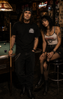 Man in black "GODS FAVORITE AZZHOE" t-shirt with woman in Misfits tank top in a dimly lit bar.