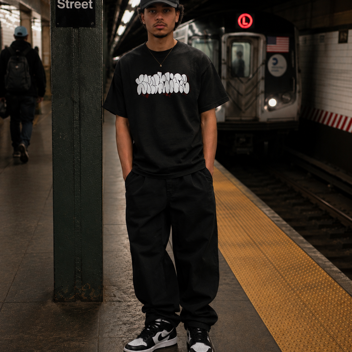 Man in black SMUTLIFE THROWIE T-shirt, black pants, and cap, standing on a subway platform.