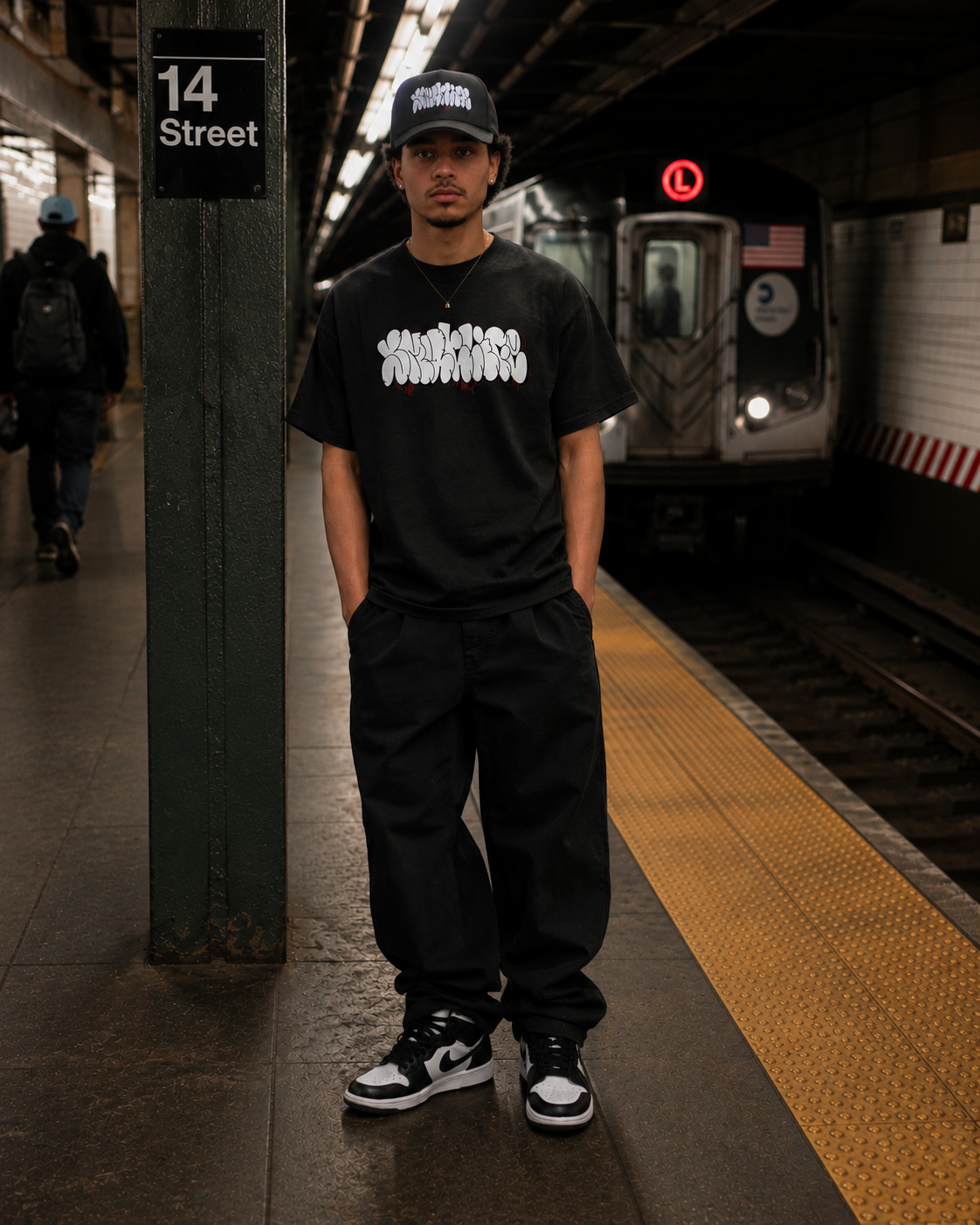 Man in black SMUTLIFE THROWIE T-shirt, black pants, and cap, standing on a subway platform.
