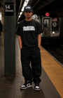 Man in black SMUTLIFE THROWIE T-shirt, black pants, and cap, standing on a subway platform.