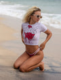 Blonde woman in white Smutlife DRIP crop top and red bikini bottom on a sandy beach.