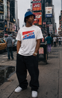 Man in white Smutlife bubble gum t-shirt, black baggy pants, blue bucket hat, and white sneakers in Times Square.