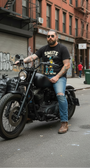Man on a black motorcycle wearing a black "SMUTS NUTS SOLD HERE" tee, ripped blue jeans, and brown boots.