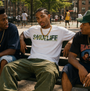 Man in white SMUTLIFE CAMO Logo Tee with gold chain, sitting on a bench outdoors.