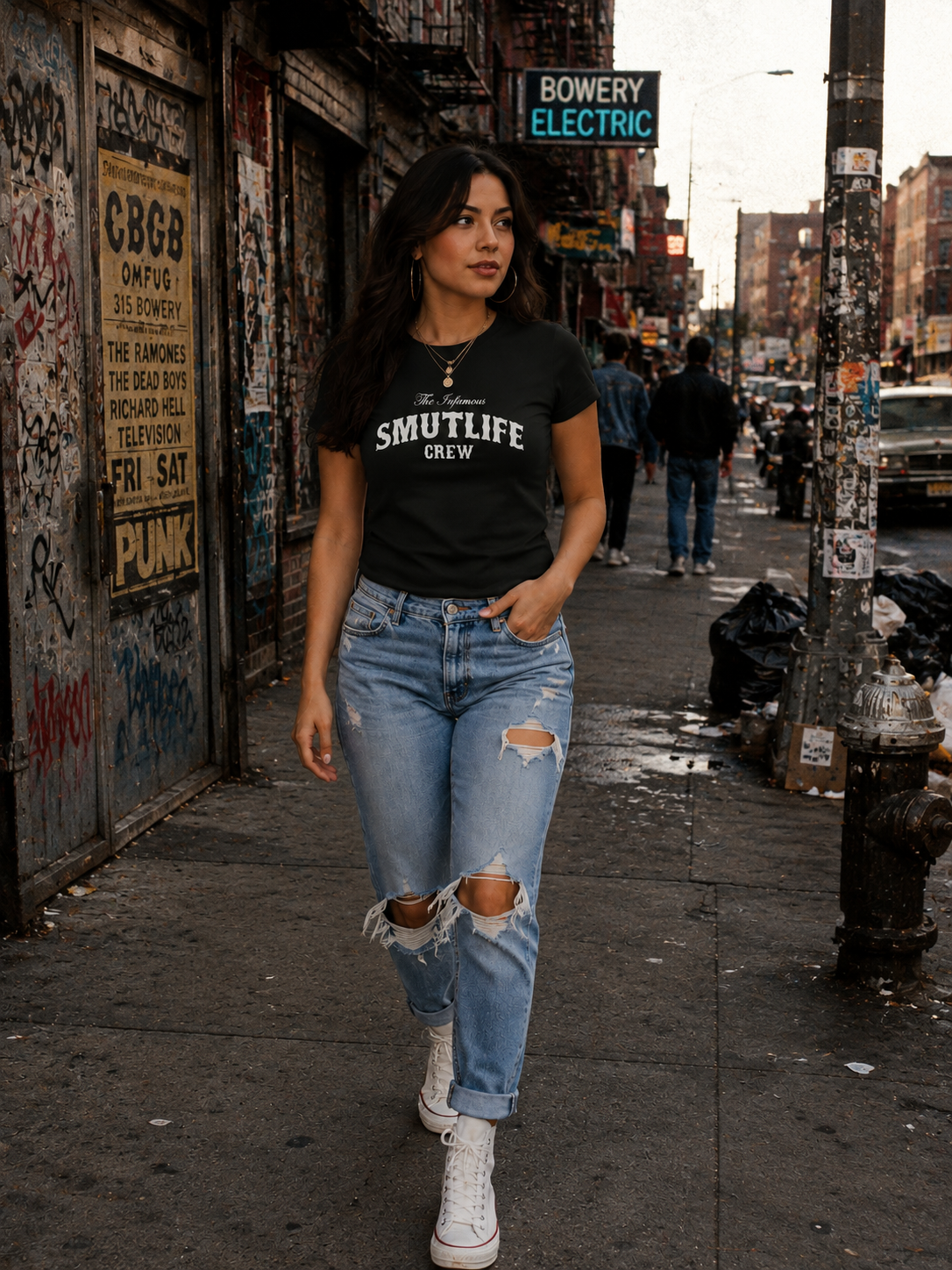 Woman in black "SMUTLIFE CREW" tee, ripped jeans, and white sneakers walking on a city street.