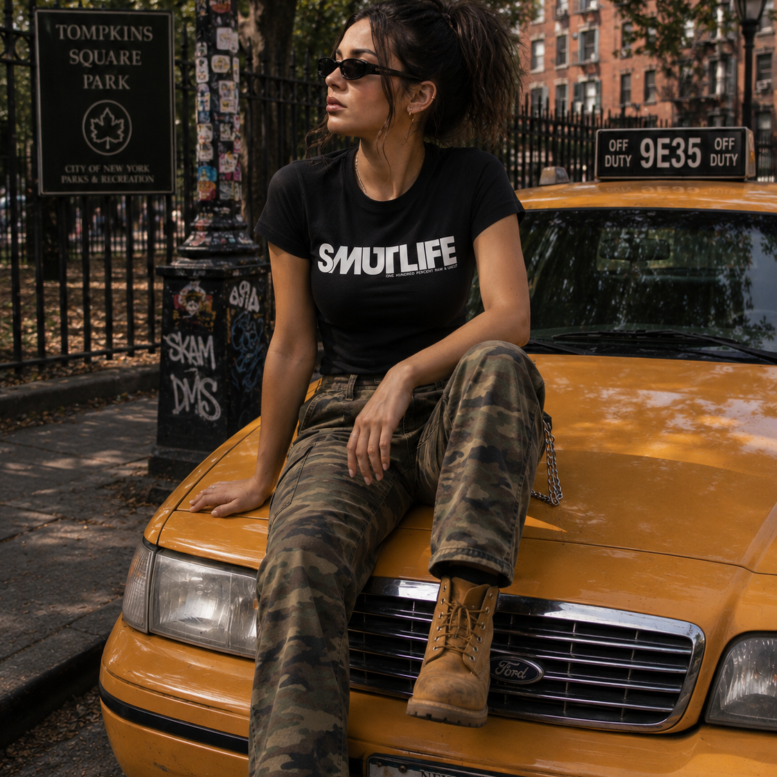 Woman in black SMUTLIFE logo tee, camo pants, and boots sitting on a yellow taxi.