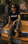 Woman in black SMUTLIFE logo tee, camo pants, and boots sitting on a yellow taxi.