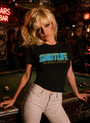 Blonde woman in a black SMUTLIFE t-shirt and light jeans, leaning on a pool table in a dimly lit bar.