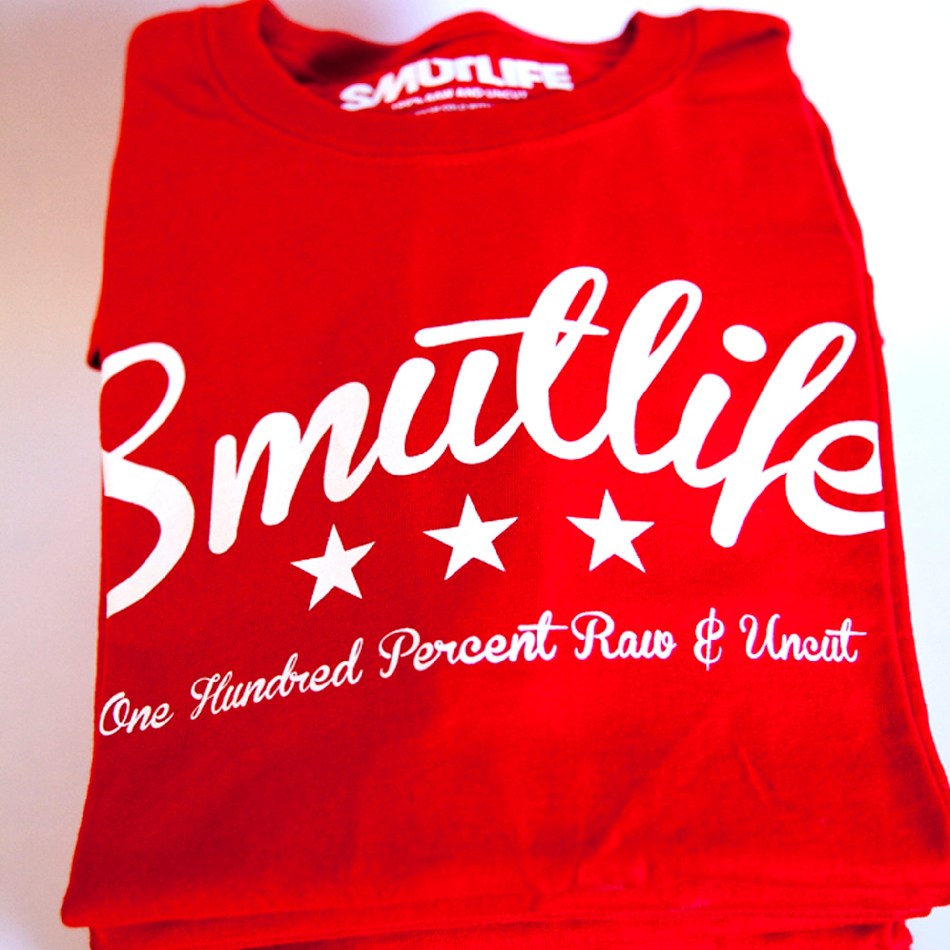 Folded red SMUTLIFE Tri-Star men's tee with white "Smutlife" text, three stars, and "One Hundred Percent Raw & Uncut" slogan.