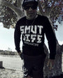 Man in black SMUTLIFE WORLDWIDE long-sleeve tee, black shorts, sunglasses, and cap, standing by a tree.