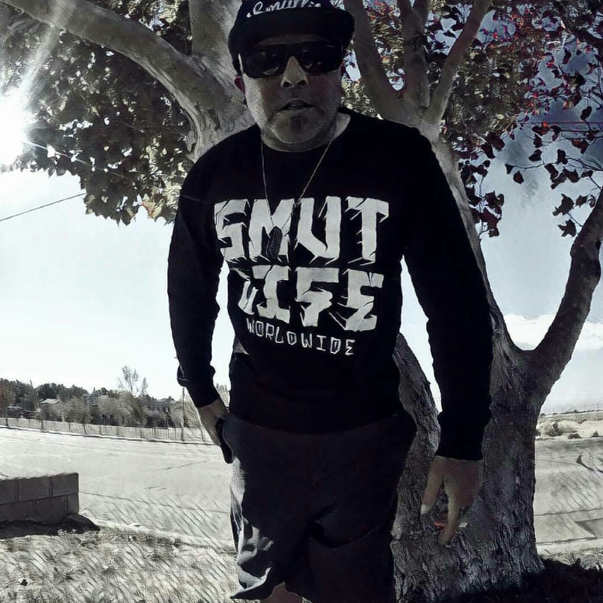Man in black SMUTLIFE WORLDWIDE long-sleeve tee, black shorts, sunglasses, and cap, standing by a tree.